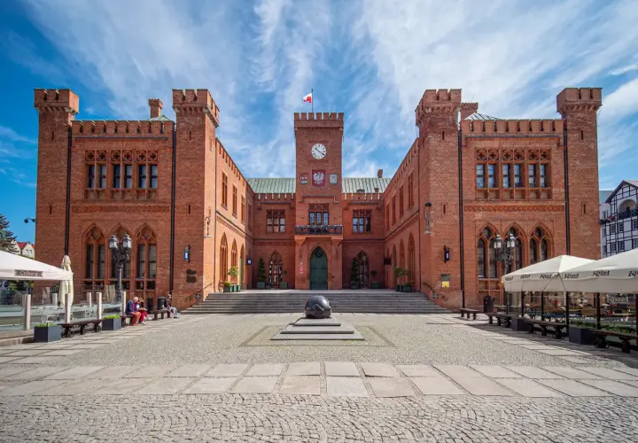 Rynek Kołobrzeg: ceglany ratusz z zegarem i flagą, przed nim kamienna rzeźba i brukowany plac.