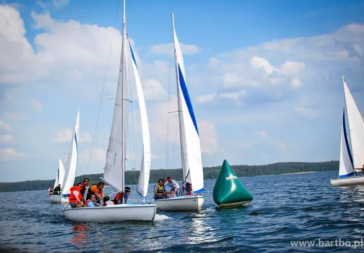 Regata żeglarska na jeziorze. Kilka łodzi z załogami ściga się, mijając zieloną bojję.