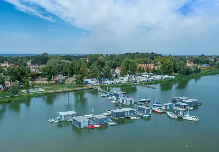 Nowoczesne houseboat'y unoszą się na spokojnej wodzie, obok kempingu z kamperami.