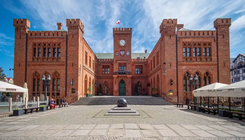 Rynek Kołobrzeg: ceglany ratusz z zegarem i flagą, przed nim kamienna rzeźba i brukowany plac.
