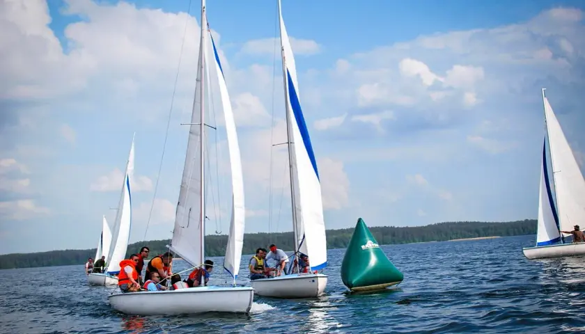Regata żeglarska na jeziorze. Kilka łodzi z załogami ściga się, mijając zieloną bojję.