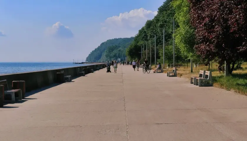 Spacerowicze i rowerzyści cieszą się słonecznym dniem na bulwarze w Gdyni. W tle widać morze i zielone wzgórza.