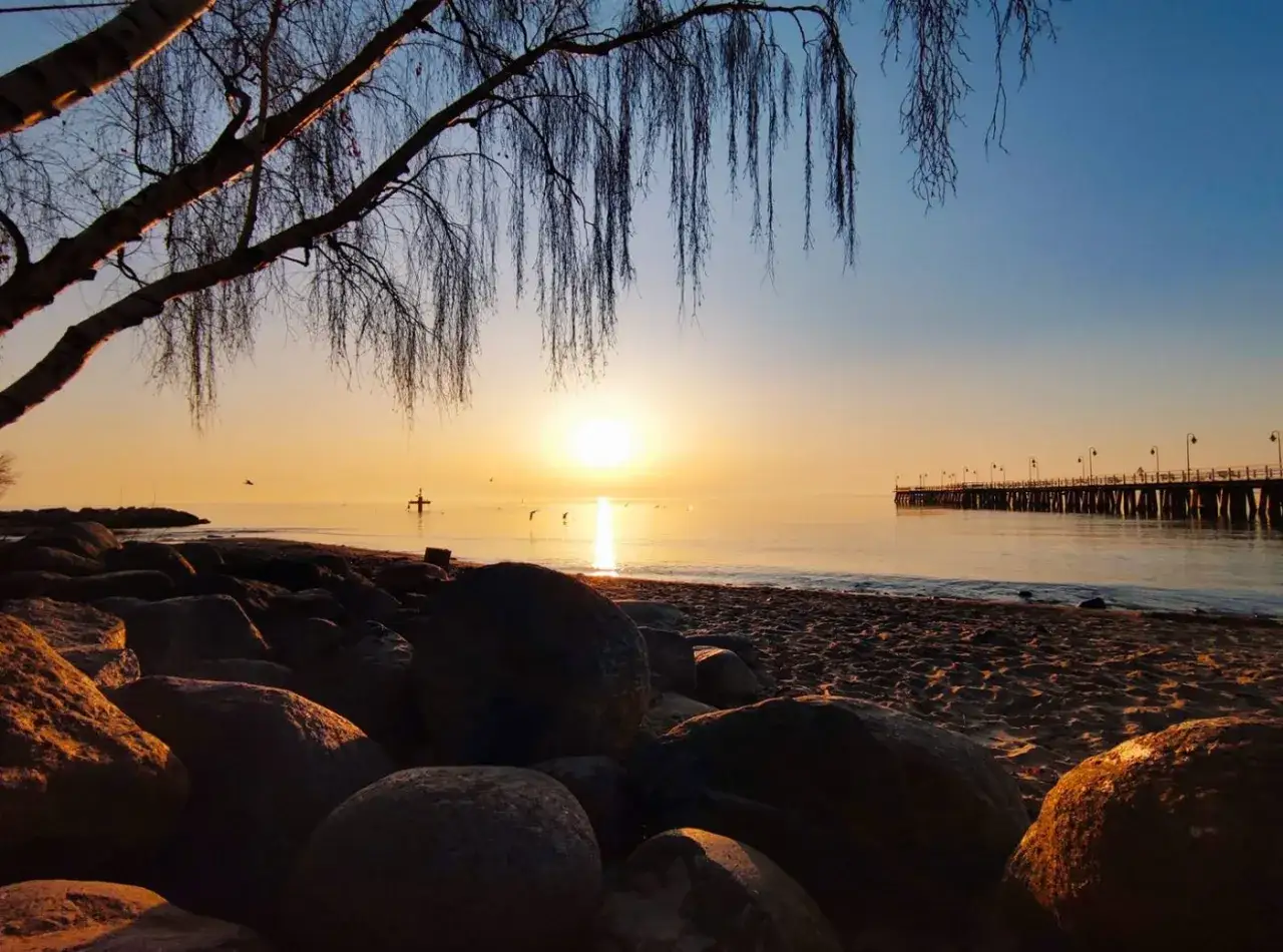 Wschód słońca w Gdyni nad spokojnym morzem, z widokiem na molo i kamienistą plażę.