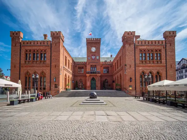 Rynek Kołobrzeg: ceglany ratusz z zegarem i flagą, przed nim kamienna rzeźba i brukowany plac.