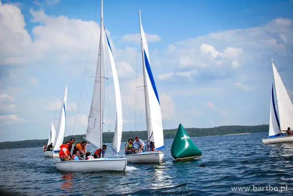 Regata żeglarska na jeziorze. Kilka łodzi z załogami ściga się, mijając zieloną bojję.